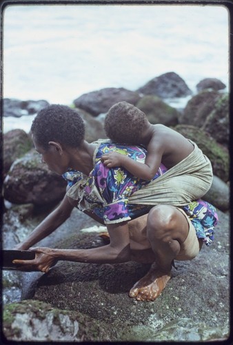 Kairiru: on rocky coastline, woman rinses a pan, small child strapped to her back