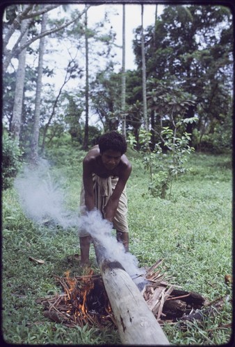 Canoe-building: man holds log over a fire, to harden the wood and smoke out termites, preparing it to be used as an outrigger on a canoe