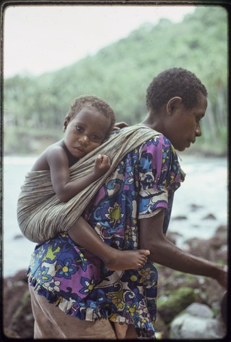 Kairiru: woman on rocky beach carries small child in a sling on her back