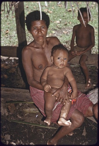 Woman holding an infant, smiling boy in background, woman's hand is tattooed