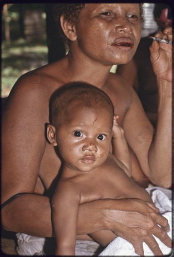 Woman with tattooed arm smokes a cigarette and holds young child, who gazes at the camera