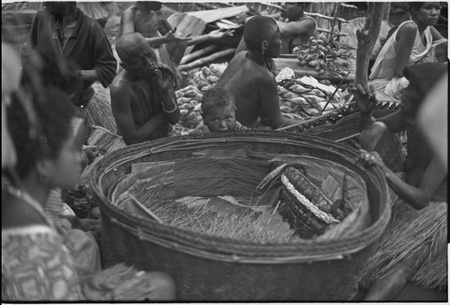 Mortuary ceremony, Omarakana: large basket of exchange valuables, banana leaf bundles stacked, woman (l) smokes