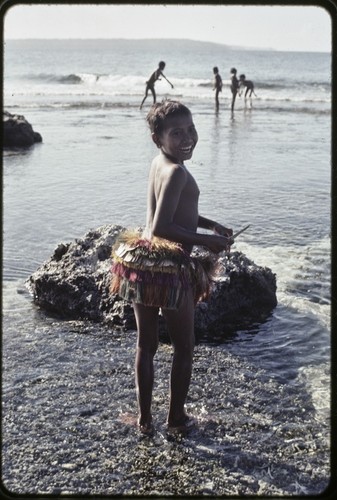 Girl in short fiber skirt walks on the fringing reef near Wawela village at low tide, other children fish on edge of coral reef in distance