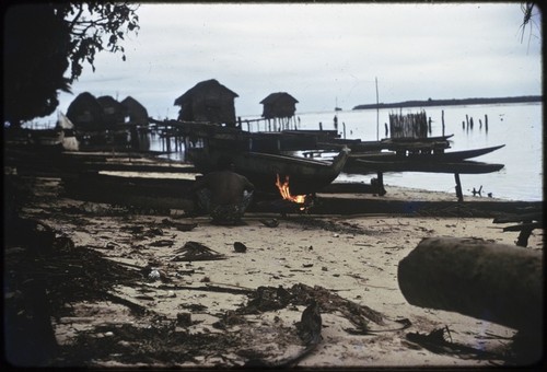 Manus: canoe-building, man heats pitch that has been applied to canoe, Pere village over-water houses in background