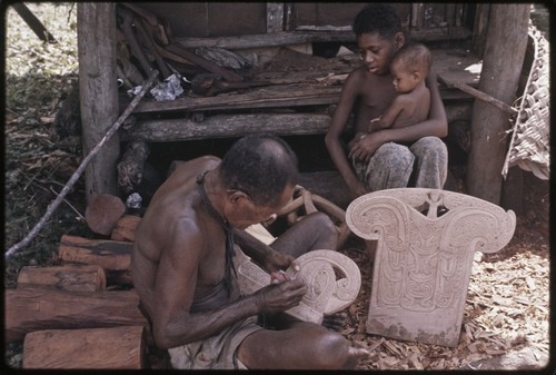 Carving: M'lapokala creates a design on canoe prowboard, next to carved splashboard, boy and infant sit nearby watching