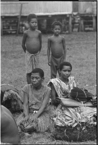 Mortuary ceremony: women sit next to basket of banana leaf bundles, woman (l) wears mourning necklace