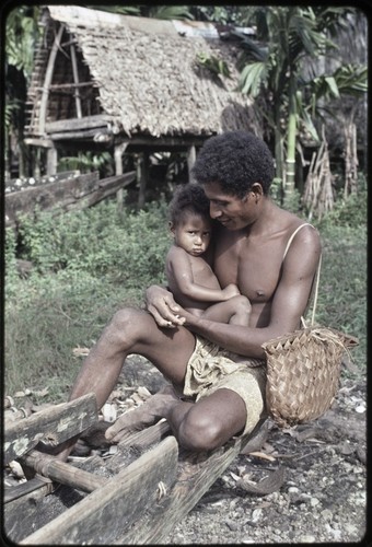 Man holding an infant, canoes and canoe house visible