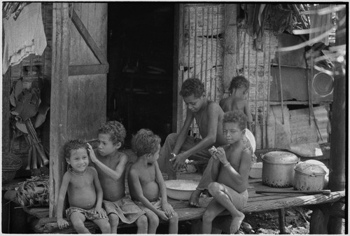 Children on a veranda, boy grates coconut into bowl, child (l) grooms younger child's hair, taro shoots (l) in background