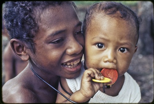 Smiling child and an infant, infant chews on a red disc commemorating Papua New Guinea's independence