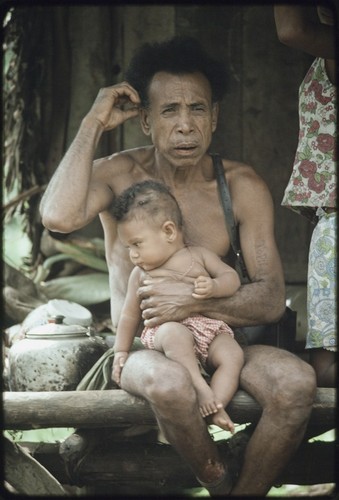 Mowaroka's father, tattoo on left arm, holds a baby