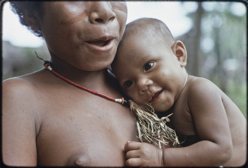 Adolescent girl, wearing shell necklace and dabs of betel nut paste on her cheeks, cuddles a baby and smiles