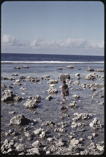 Girl in short fiber skirt walks on a reef near Wawela village at low tide, waves break on edge of coral reef in distance