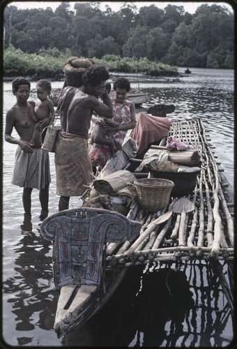 Canoes: man places basket of yams and other items onto canoe for coastal transport, women holding infants stand nearby