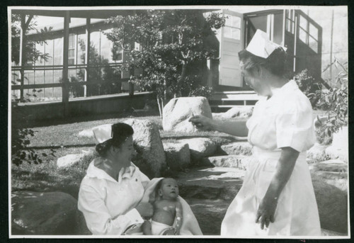 Photograph of Irene Gavigan, R.N., holding a newborn while another nurse attracts his attention in front of the Manzanar hospital