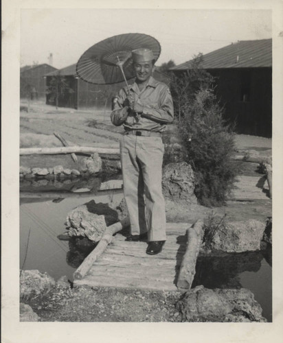 Mitsuru Yagura holding an umbrella on a wooden footbridge over a pond at Poston incarceration camp