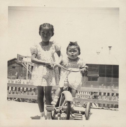 Adolescent girl standing next to toddler on a tricycle at Poston incarceration camp