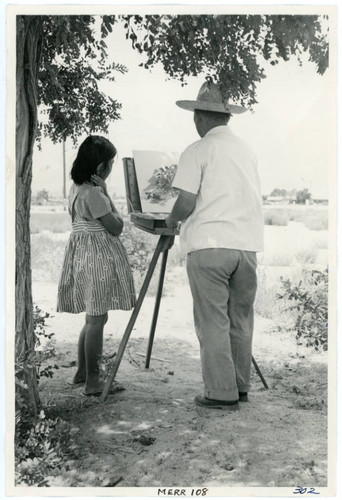 Photograph of an artist painting at an easel while a young girl watches