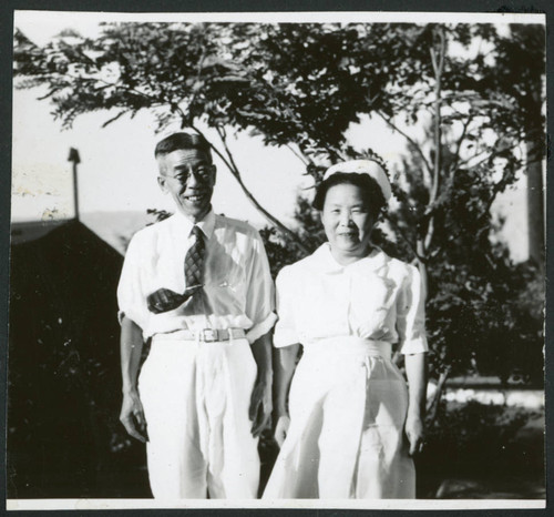 Photograph of a couple standing in front of trees and buildings at Manzanar