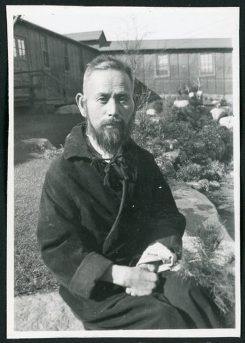 Photograph of a man in a robe sitting on a rock wall in front of the Manzanar hospital