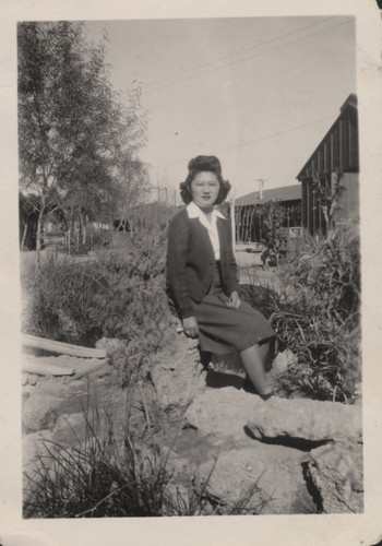 Young woman in cardigan sweater sits at a pond at Poston incarceration camp