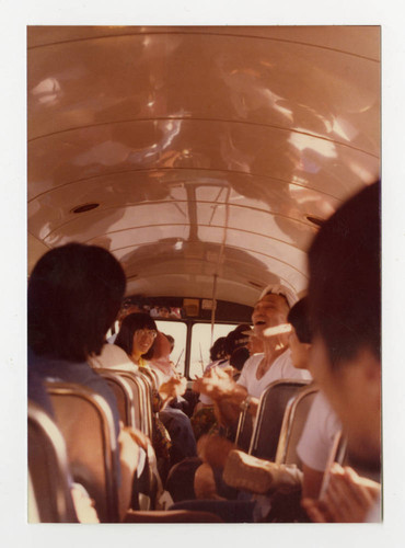 A photo of people sitting and talking on the a bus to or from the 10th annual Manzanar Pilgrimage. Sansei Tamiko Hirano faces the camera