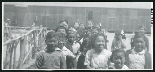 Photograph of children at Manzanar standing next to a fence and in front of a barracks