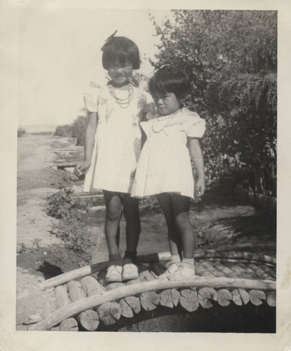 Two little girls wearing necklaces stand on a footbridge over water at Poston incarceration camp