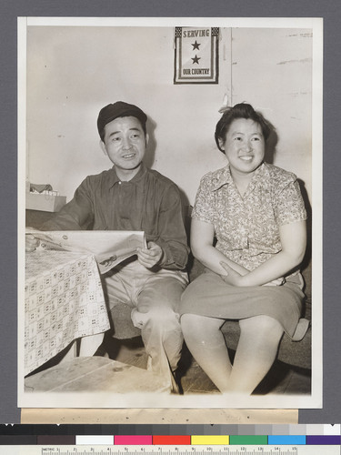 Tom and Susie Sugimoto, man and wife, are seated in their apartment at the Tulelake (correct) Japanese Relocation Center in Northern California. The service flag over their heads is for brothers of Susie who lived with them until they entered the armed service