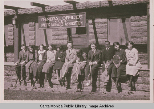 A group of men and women with tennis racquets sit in front of a building identified as "General Offices - Pacific Palisades Association."