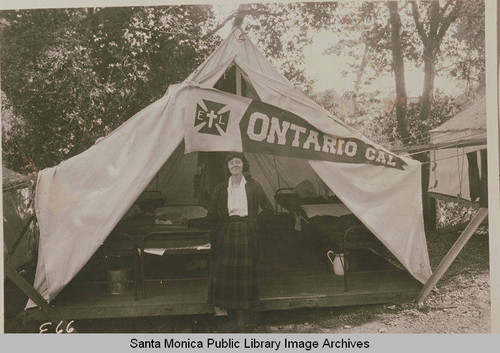 A woman stands in front of a tent with a banner labeled "Ontario CA," at the Institute Camp, Temescal Canyon, Calif