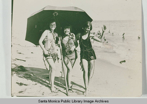 Three women in bathing suits draped in seaweed posing under an umbrella on the beach