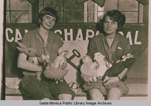 Two young women with an assortment of childrens' toys sit in front of banner "Camp Chaparral," Pacific Palisades, Calif