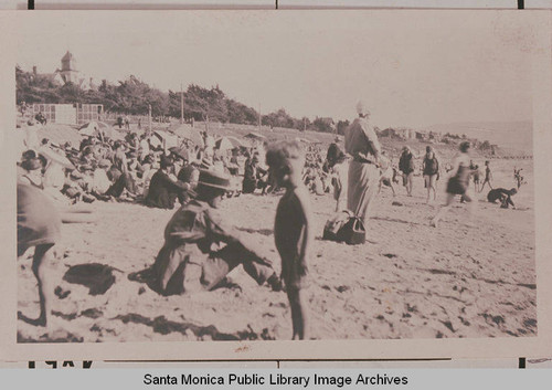 A crowded day at the beach in Long Beach, Calif. showing children in the sand as adults sit in the sun or beneath umbrellas facing the water