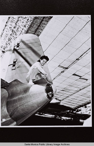 Douglas Aircraft Company public relations shot of a woman standing on the tail section of an airplane using a screwdriver under camouflage, World War II