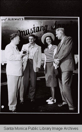 Actress Dorothy Malone, KMPC reporter and the "Mustang Special" airplane (Southern Methodist University Mustangs) on the tarmac near Douglas Aircraft Company, Santa Monica with the slogan "SMU Beat UCLA."