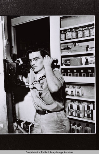 Douglas Aircraft Company employee in front of a shelf of personal supplies answering a wall mounted telephone during World War II