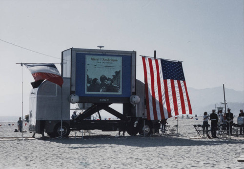 Large screen flanked by American and French flags showing 'Merci l'Amerique' during the reenactment of D-Day landing, Santa Monica, Calif