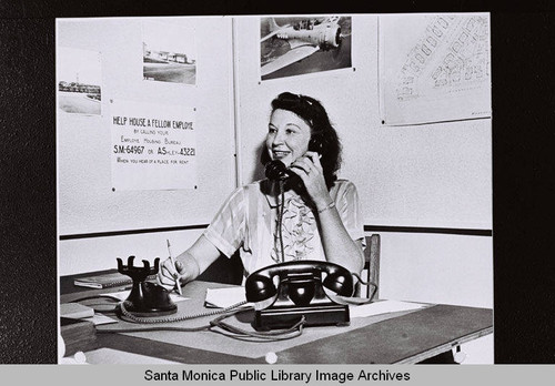 Douglas Aircraft Company Housing Bureau clerk answering telephones by posted sign "Help House a fellow Employe by calling your Employe Housing Bureau" during World War II