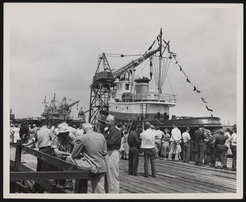 Crowd stands on dock by tugboat