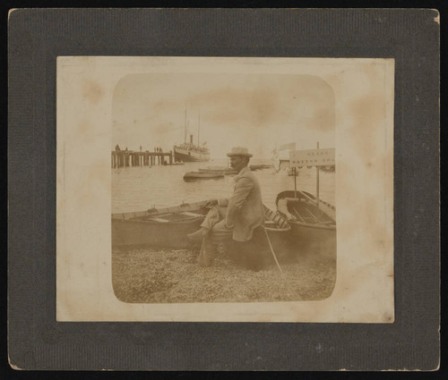 Man in white suit and hat sits on boat at Avalon Harbor, front of photograph