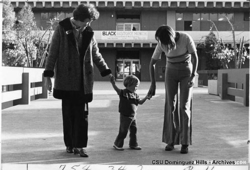 Two people help child walk toward the Library