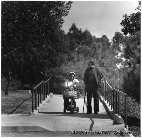 Older student in electric cart walks with student with cane