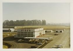 Sonoma County Airport main hanger with "Let's fly Piper" sign mounted at the top side of the building, 1973