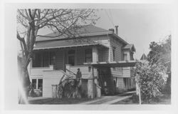 Unidentified woman standing on the steps of a two-story house with a large arbor over the side driveway, 1960s or 1970s