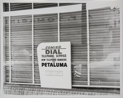 Store window sign for Pacific Telephone's "new" Dial Telephone Service with new telephone numbers