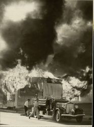 Petaluma Fire Department firefighters at their pumper truck next to a commercial building engulfed in flames, 1950s