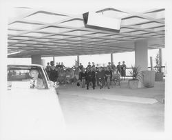 Man drives a white Plymouth Fury while a model displays a black dress behind him in a fashion show at dedication of parking garage at 3rd and D, Santa Rosa, California, 1964