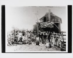 Hop pickers wearing hop garlands on their hats gathered in front of a barn