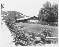 Single-story suburban home with a wide, low overhang, surrounded by a large lawn edged with a split-rail fence and located below an oak-covered hillside in an unidentified location in Sonoma County, California, 1950s or 1960s