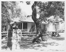 Bell mounted on a stone column, possibly in a park, with a man re-roofing a single story house with wooden shingles in the background, located in an unidentified location in Sonoma County, California, 1960s or 1970s